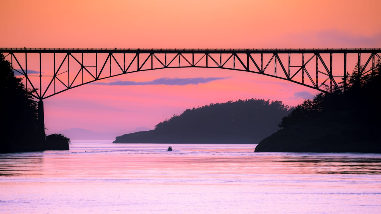 A boat passing under a bridge over a body of water at sunset. The bridge is made of concrete and has a cable-stayed design.