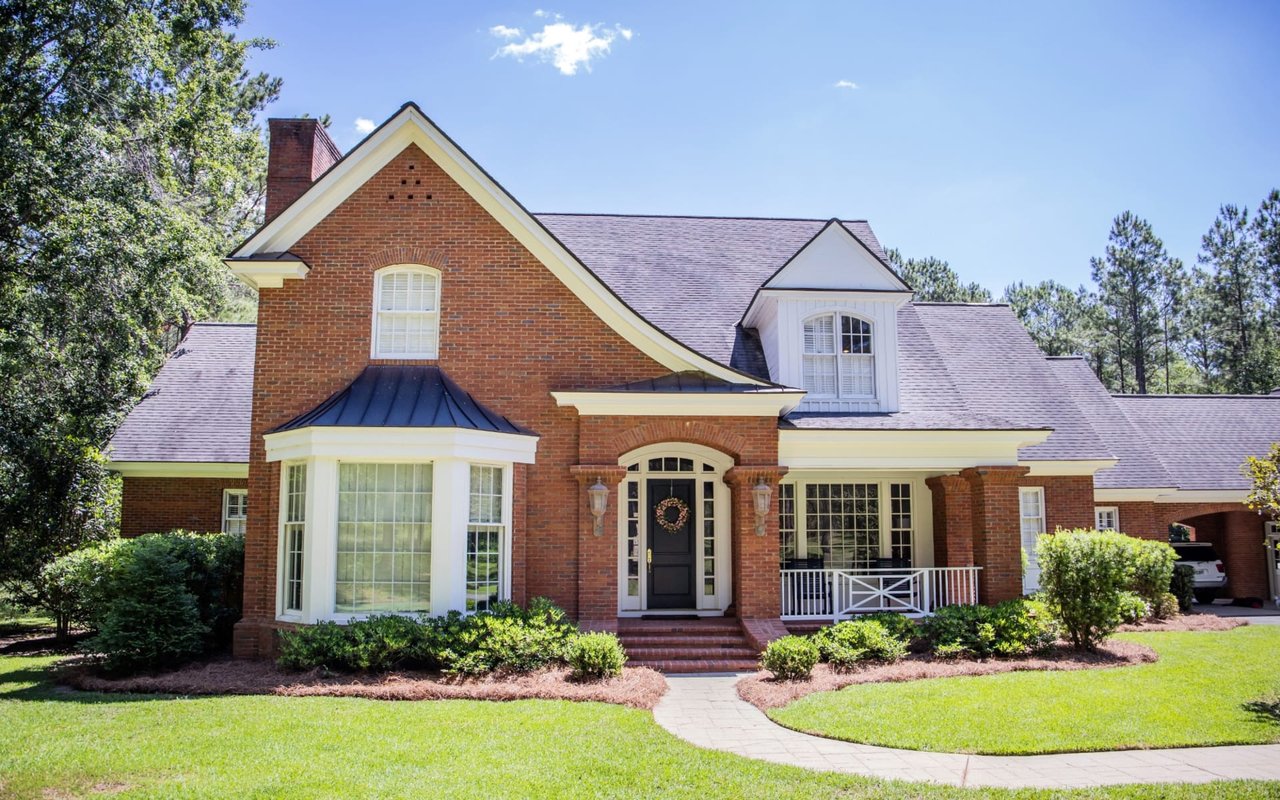 A charming brick house with white trim and a black door sits amidst lush greenery.