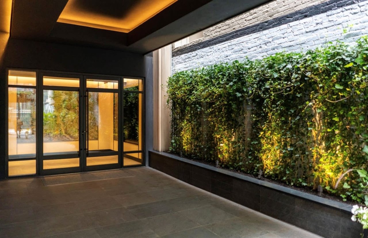 Sheltered entrance courtyard with glass doors, a leafy green wall, and warm ceiling lights.