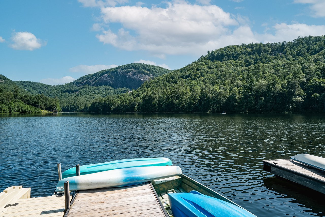A serene lake nestled between green, forested hills under a partly cloudy sky. The foreground features a wooden dock with kayaks and boats moored. Real estate agents might note its peaceful and picturesque setting, radiating tranquility and natural beauty—an idyllic spot for potential buyers.