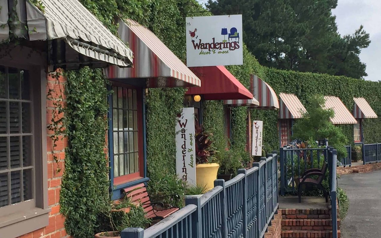 A charming street with storefronts and outdoor seating areas. The area appears to be part of a quaint town or community, with ivy-covered buildings and decorative awnings.