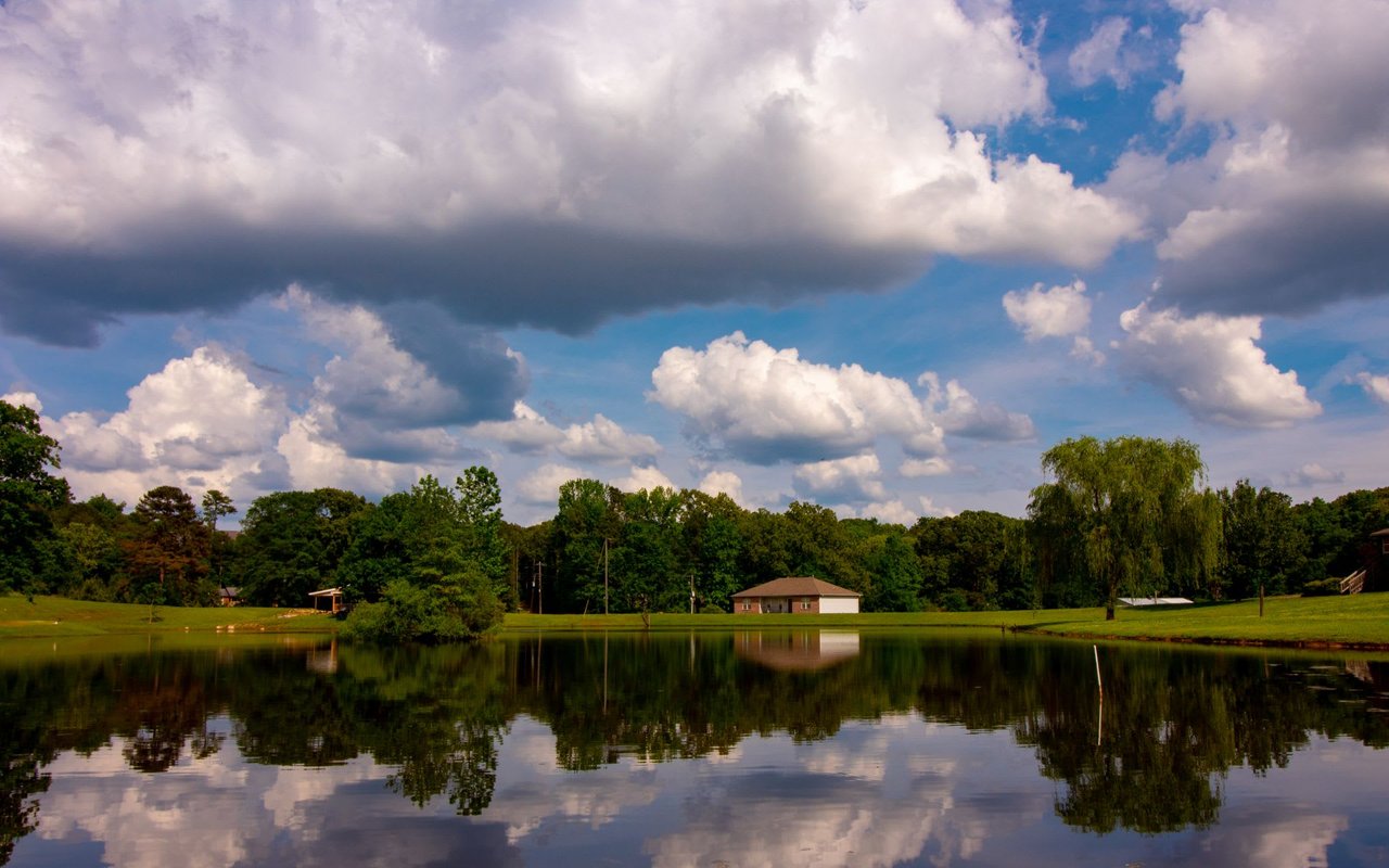 Lake scene with clear reflections of trees and a house under a cloudy blue sky.