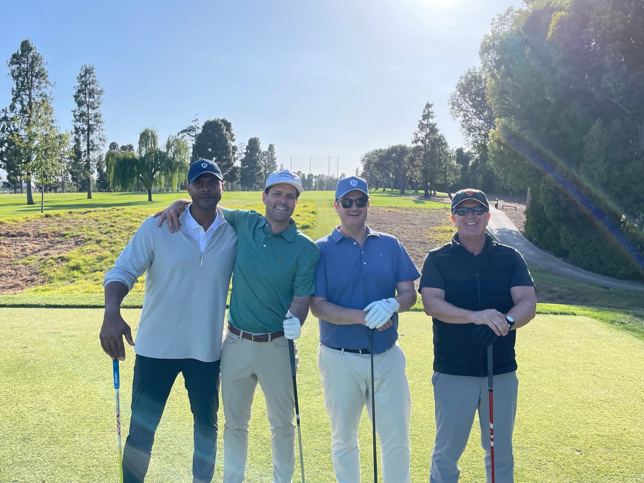  Four men are standing on a golf course under a clear, bright sky. 