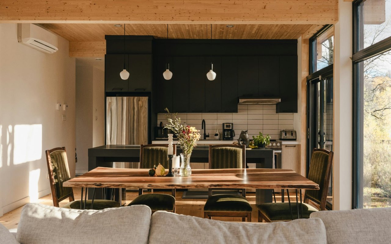 A modern dining area and kitchen with a wooden table, chairs, hardwood floor, white walls, and a large backyard-view window.