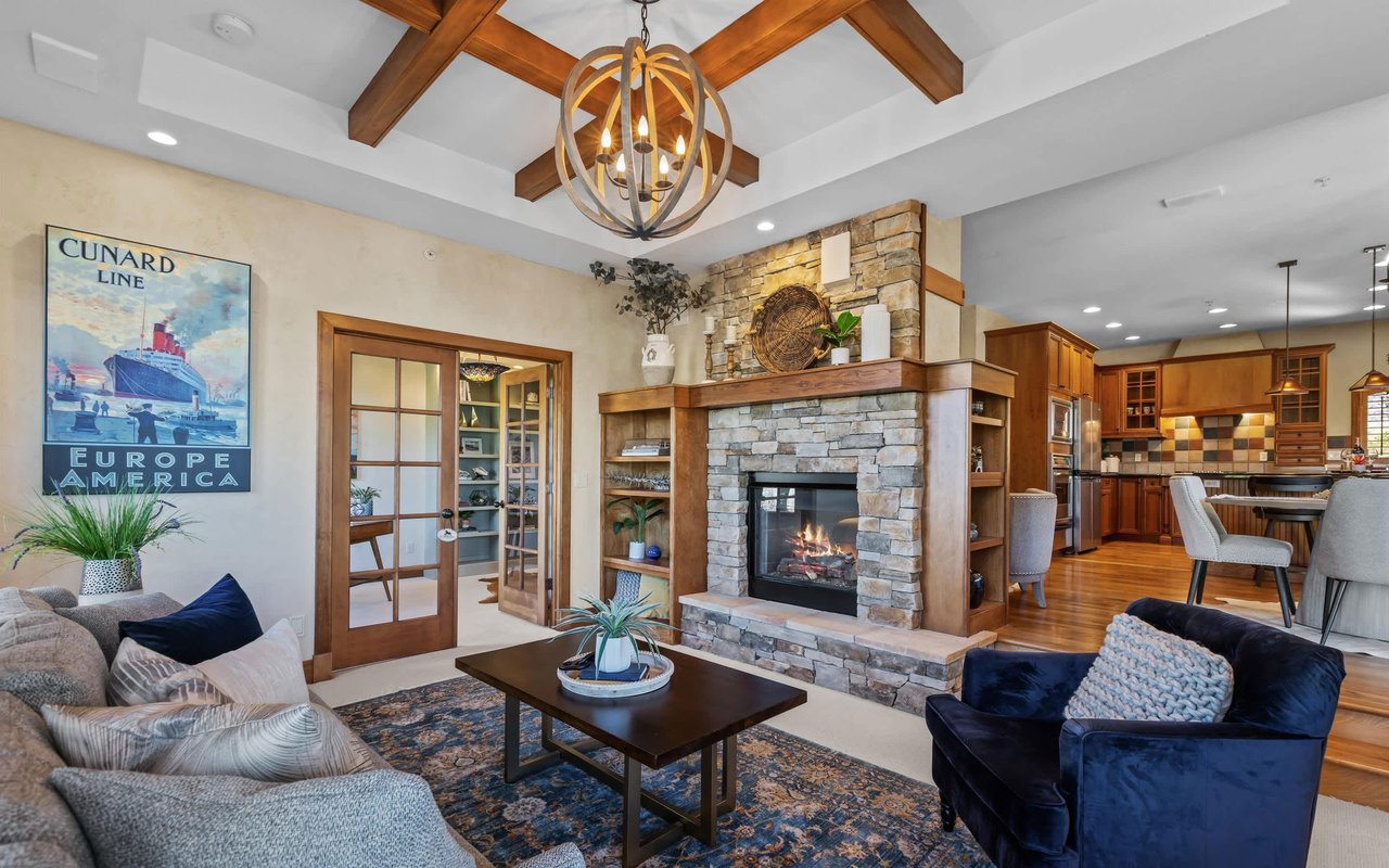 Wide-angle view of the living room, dining room and kitchen, as well as the French doors leading into the library. Points of interest include the coffered ceiling and double-sided fireplace.