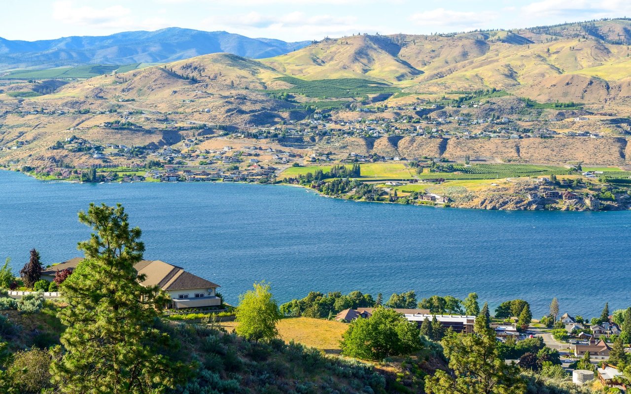 A large house with a light-colored roof is nestled among green trees and brush on a hillside overlooking the lake.