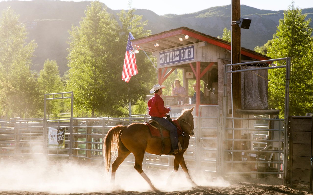 Cowboy on horseback entering the Snowmass Rodeo arena with American flag and mountain scenery in the background, capturing the authentic Western heritage experience available in Snowmass Village during summer evenings.