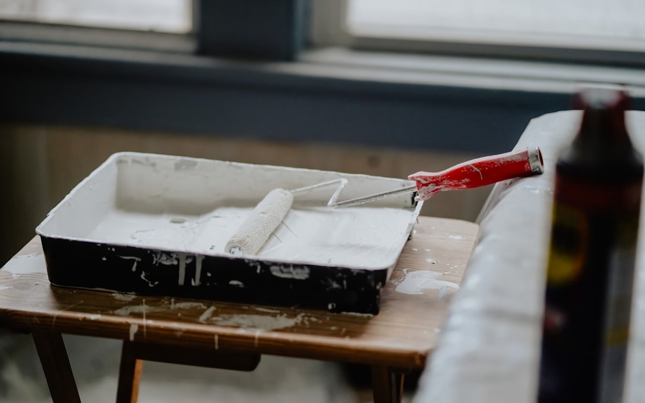  A white paint roller and a black paint tray filled with white paint are sitting on a light wooden table, which is covered in paint splatters.