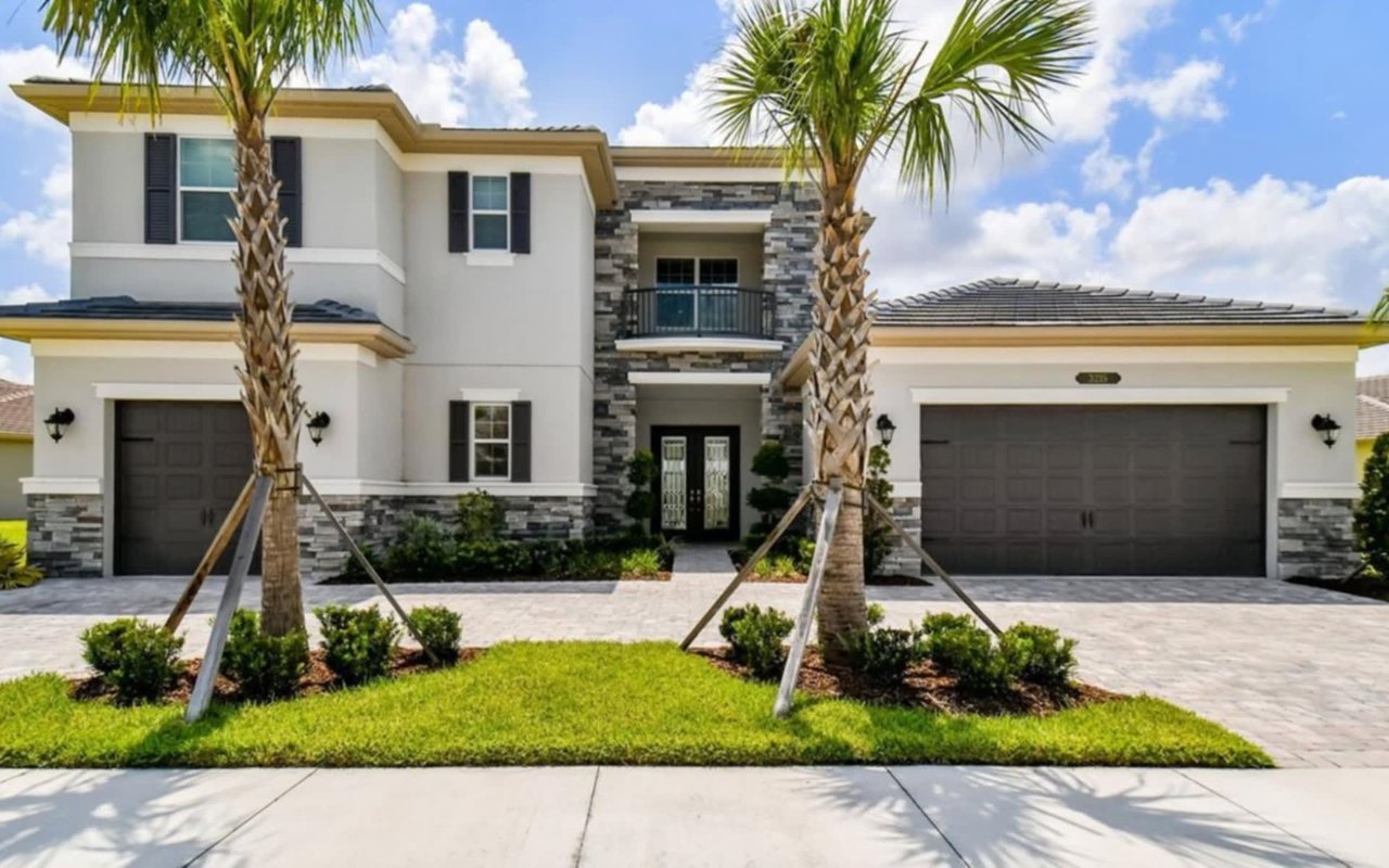 A modern two-story house with a white stucco exterior, two-car garage doors, tile roofs, and palm trees in the front yard.