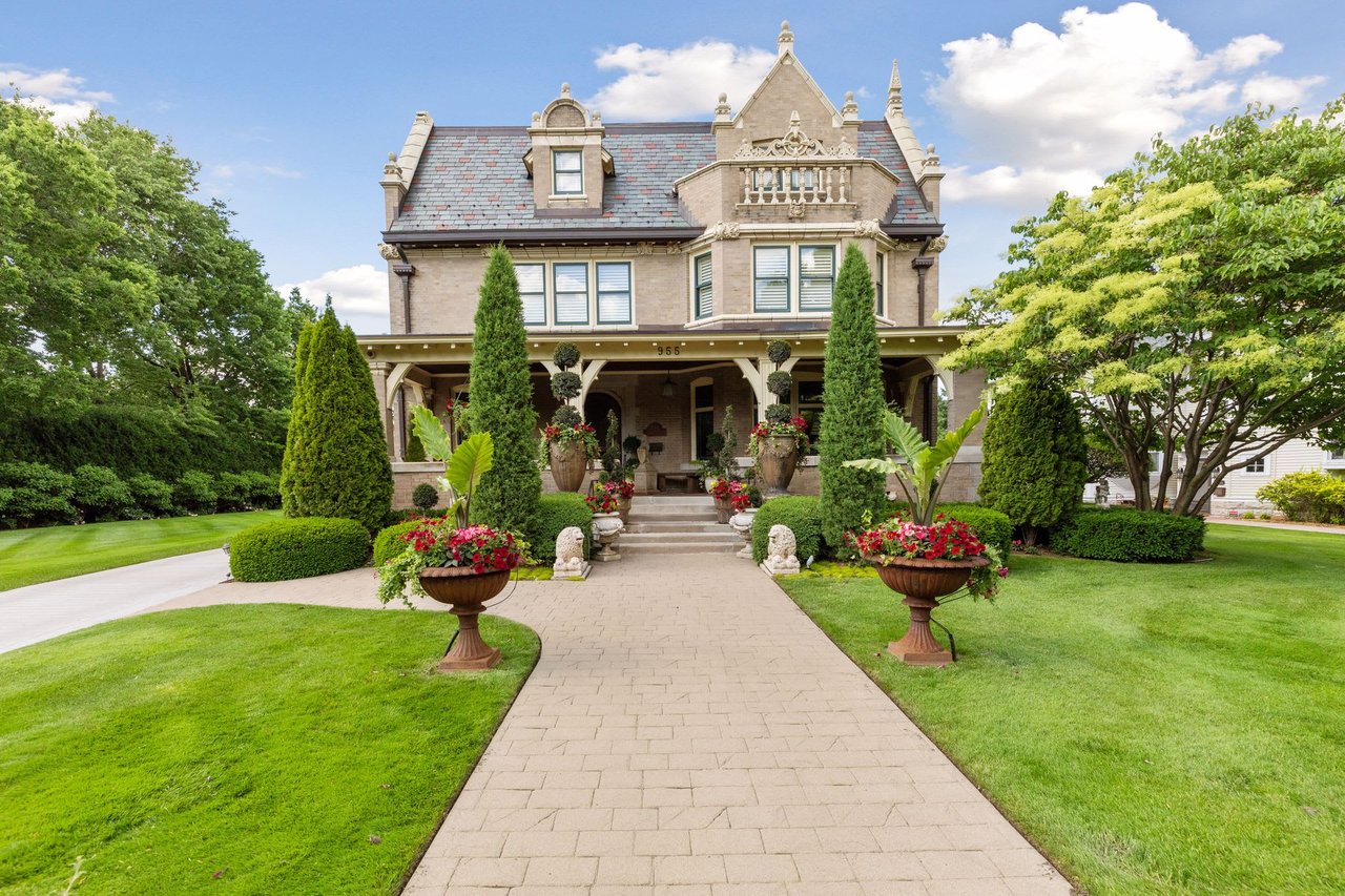 A house with a slate roof, large porch, brick walkway leading to the house surrounded by trees and bushes.