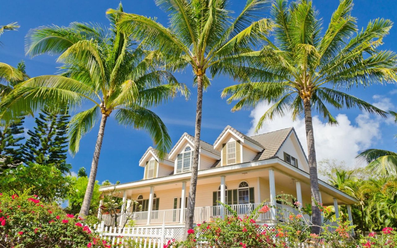 A white house with a red tile roof, black shutters, and a wrought-iron fence surrounded by lush greenery.