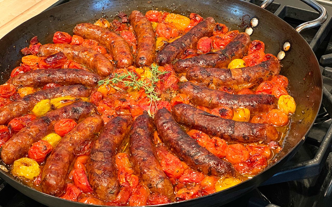 Italian sausages cook in a skillet with roasted red and yellow tomatoes and fresh herbs.