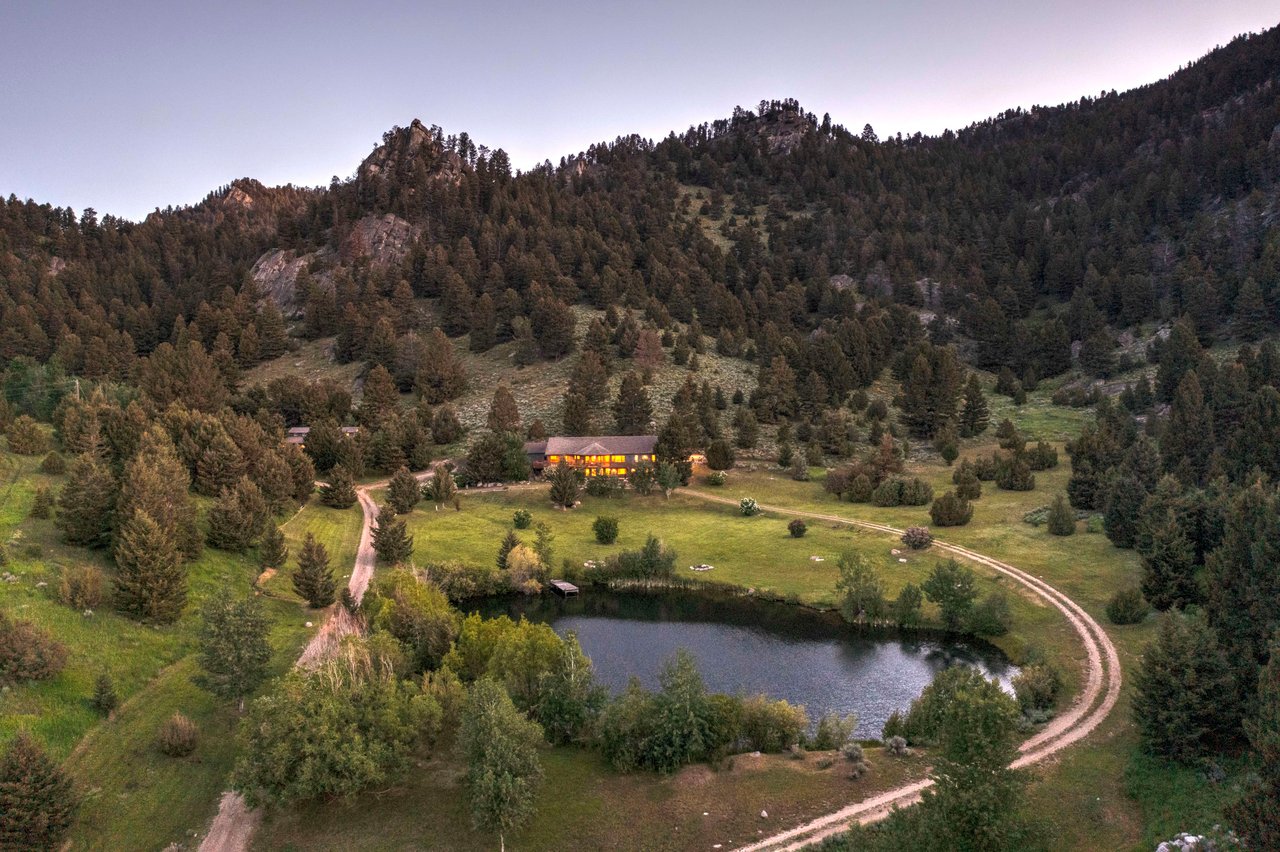  An aerial view of a secluded mountain retreat at dusk, showing a large lodge or house with warm lights glowing from its windows.