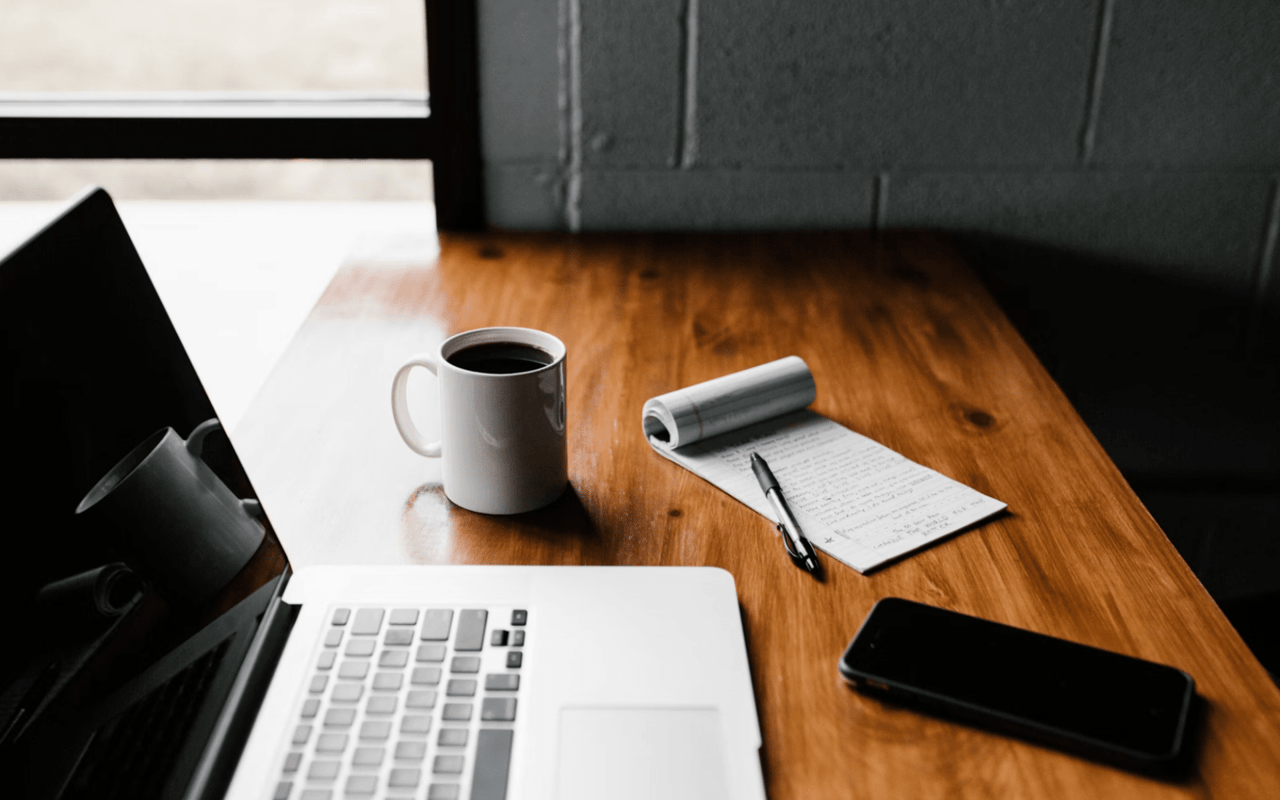 A workspace with a laptop, a mug of coffee, a notepad with a pen, and a smartphone on a wooden desk