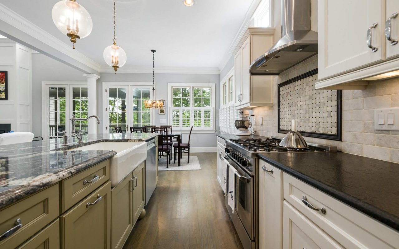  A well-appointed kitchen features a long island with a dark granite countertop and a white farmhouse sink in the foreground, flanked by light olive-green cabinets with silver handles.