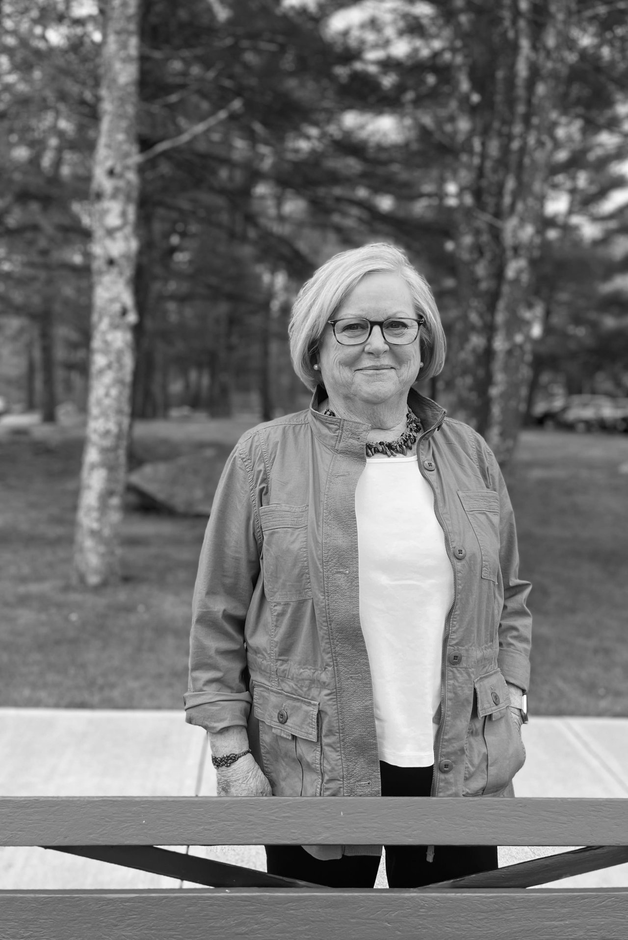 Mary Anne Condon wearing glasses and a light jacket stands outdoors in a wooded area, smiling at the camera. The image is in black and white.