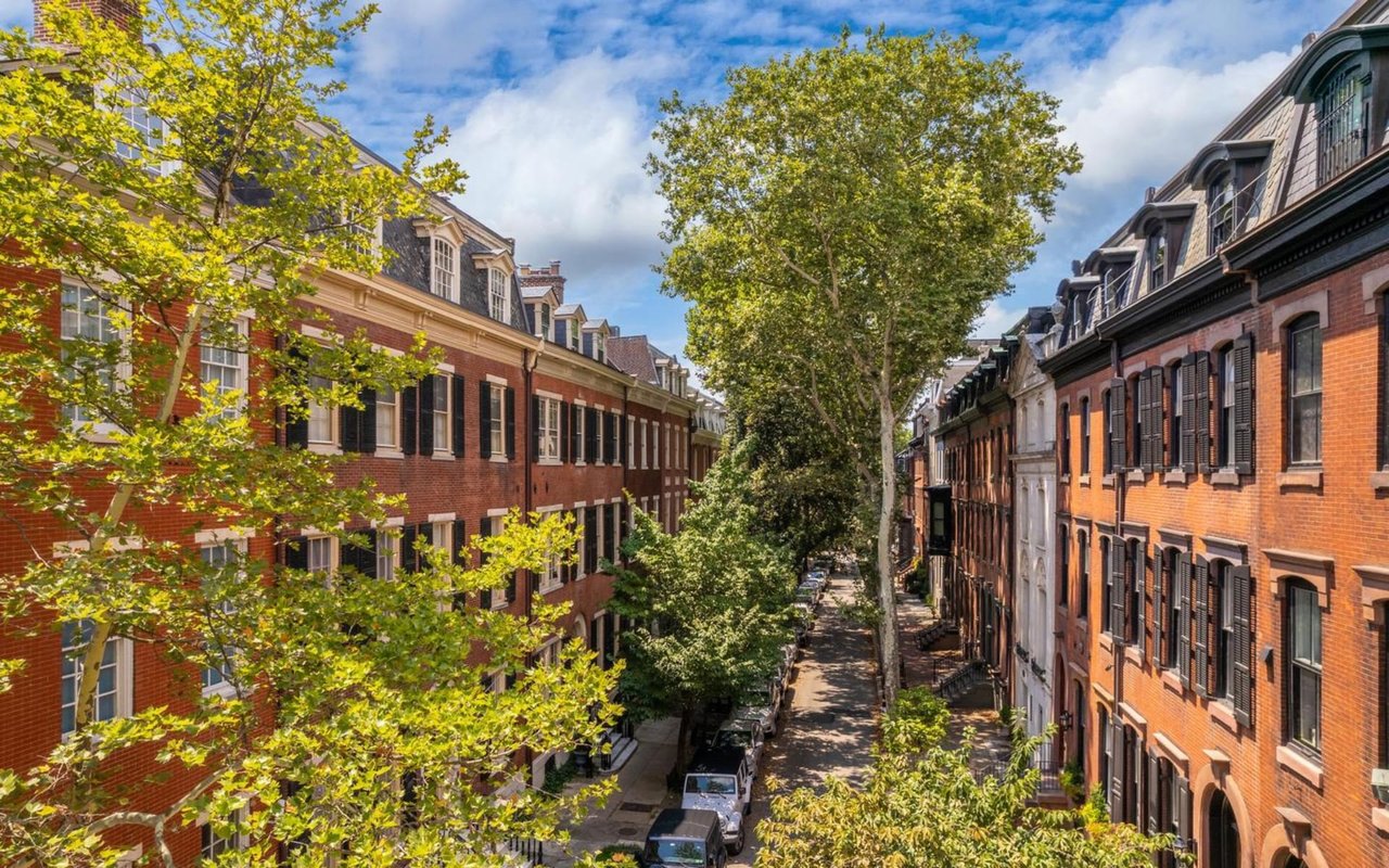 A vibrant street lined with historic red-brick row houses, framed by tall green trees.