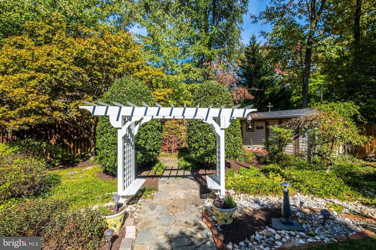 Backyard stone path beneath a white pergola, surrounded by mature trees and garden beds.