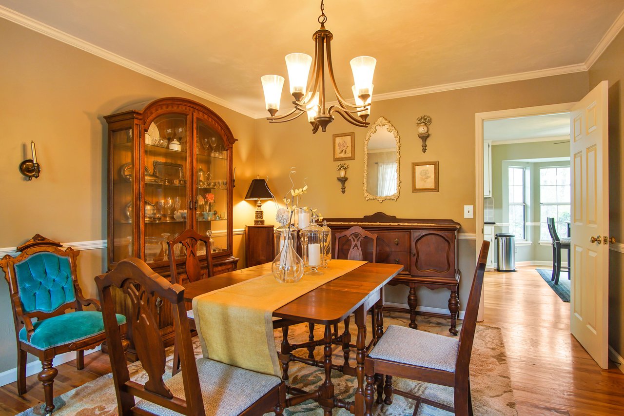 The room features a wooden dining table, chairs, a chandelier, and is decorated with paintings and a china cabinet.