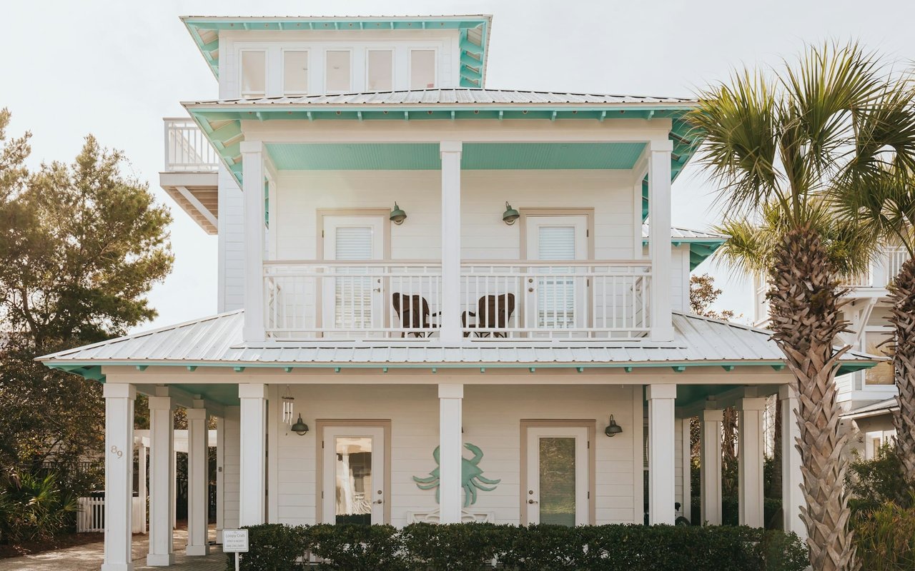 A white two-story beach house with a teal-colored roof and a large front porch. 