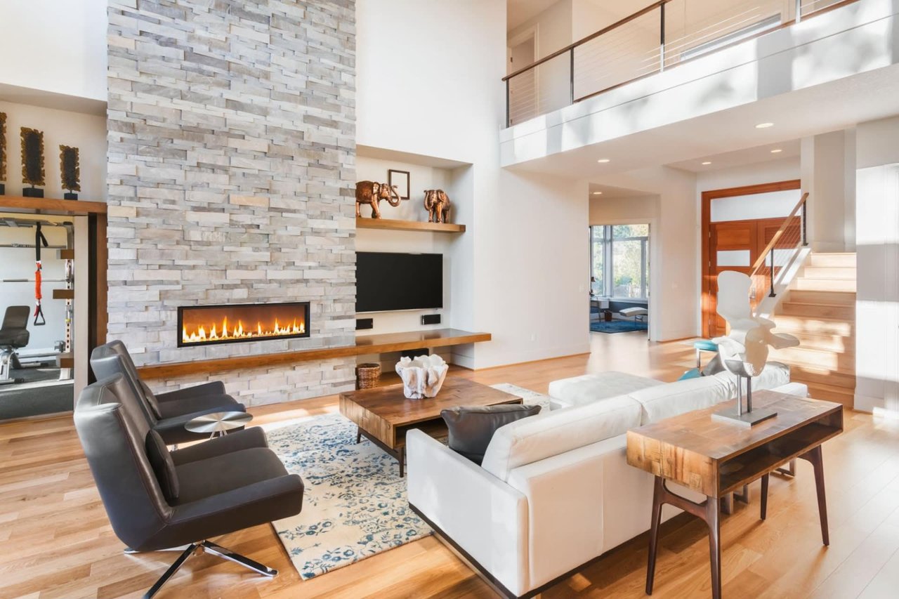  A living room with a stone fireplace, hardwood floors, windows, a white sofa, armchairs, a patterned rug, and a mounted TV.