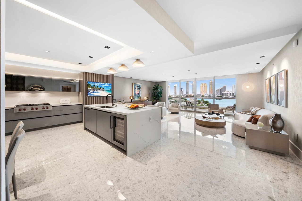 Modern kitchen/living area with gray cabinets, large island, city view, and sleek furnishings, featuring upscale appliances.