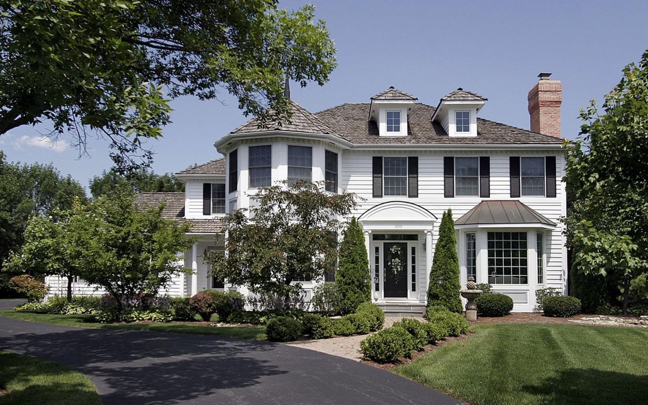 A large white house with black shutters, a gray roof, a turret, a porch, and a manicured lawn.