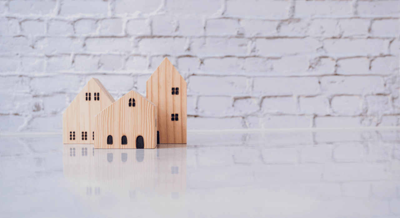 Three wooden toy houses reflected on a white surface, with a brick wall in the background.