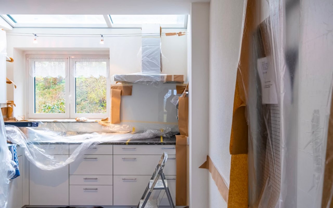 A kitchen undergoing renovation with plastic-covered cabinets, an outdoor-view window, and a doorway leading to another room.