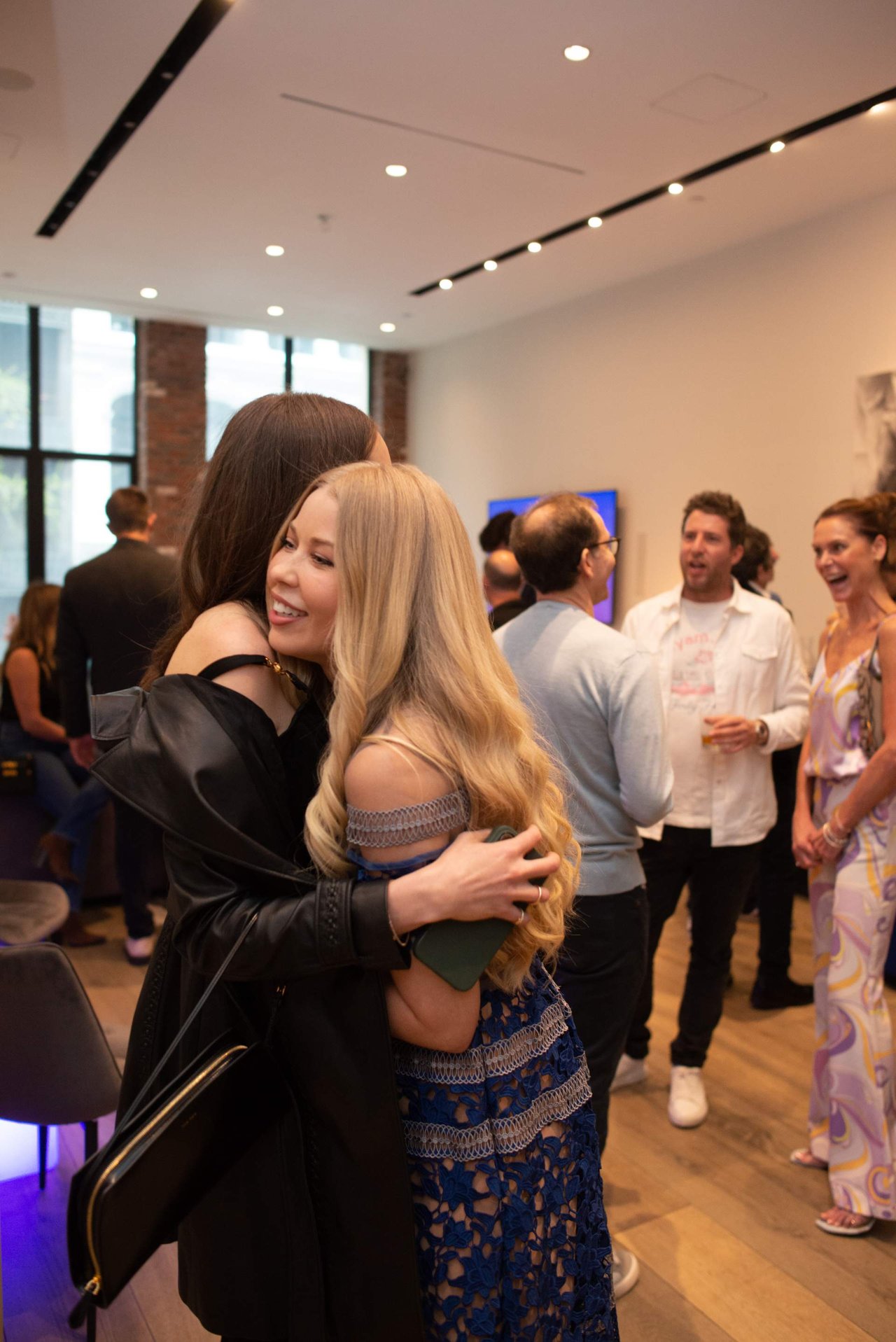 Two women embrace as guests mingle at an indoor event.