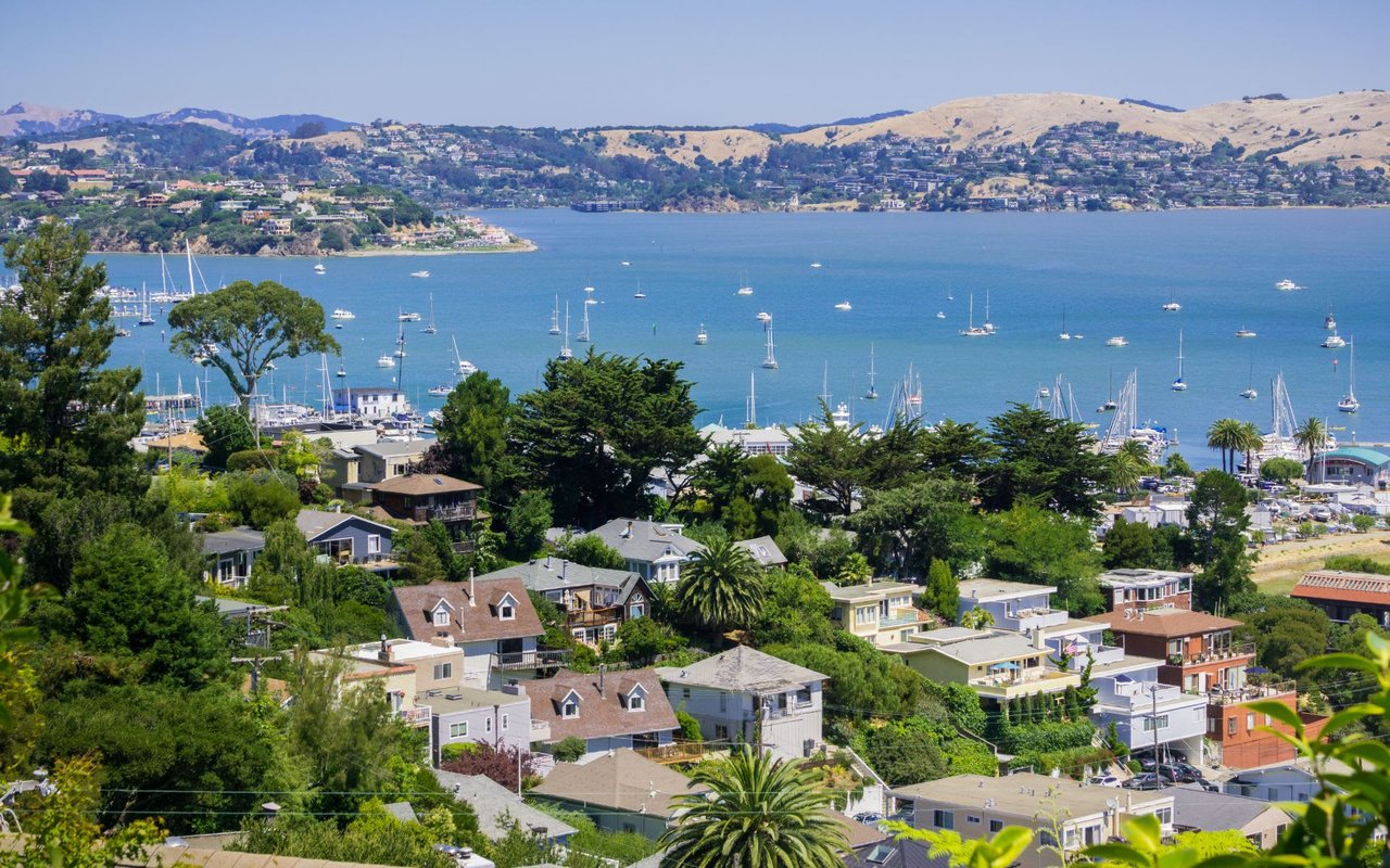 A hillside town with colorful houses overlooks a bay filled with sailboats under a clear blue sky.