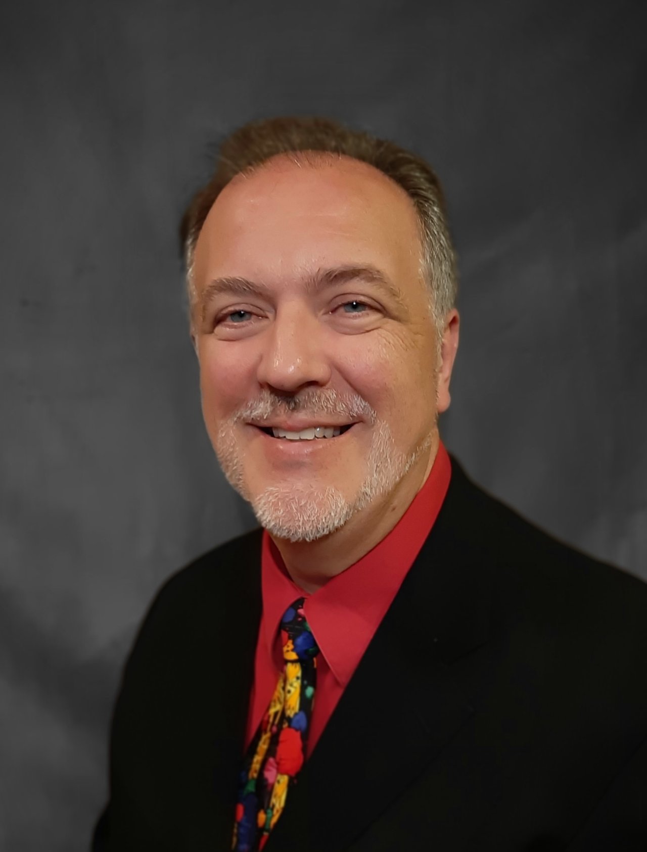 Rick VanCuren smiling in a black jacket, red shirt, and colorful patterned tie.