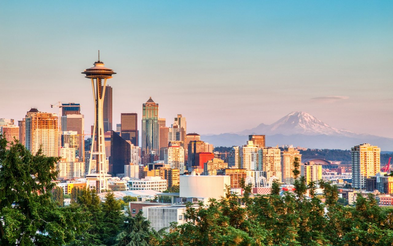 A city skyline with skyscrapers and there is a snow-capped mountain peak and a partly cloudy blue sky in the background.