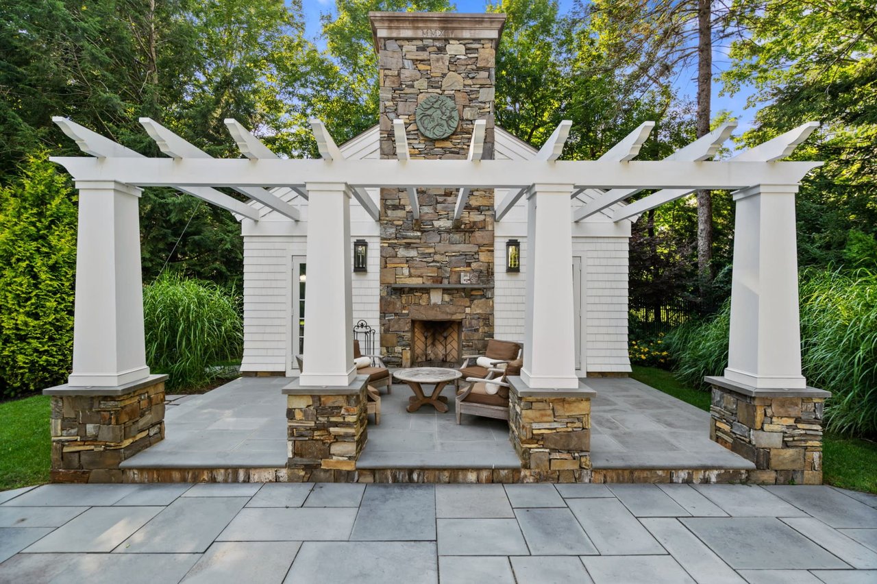  A luxurious outdoor living space featuring a white pergola with thick posts, some of which are clad in stone, over a stone patio.