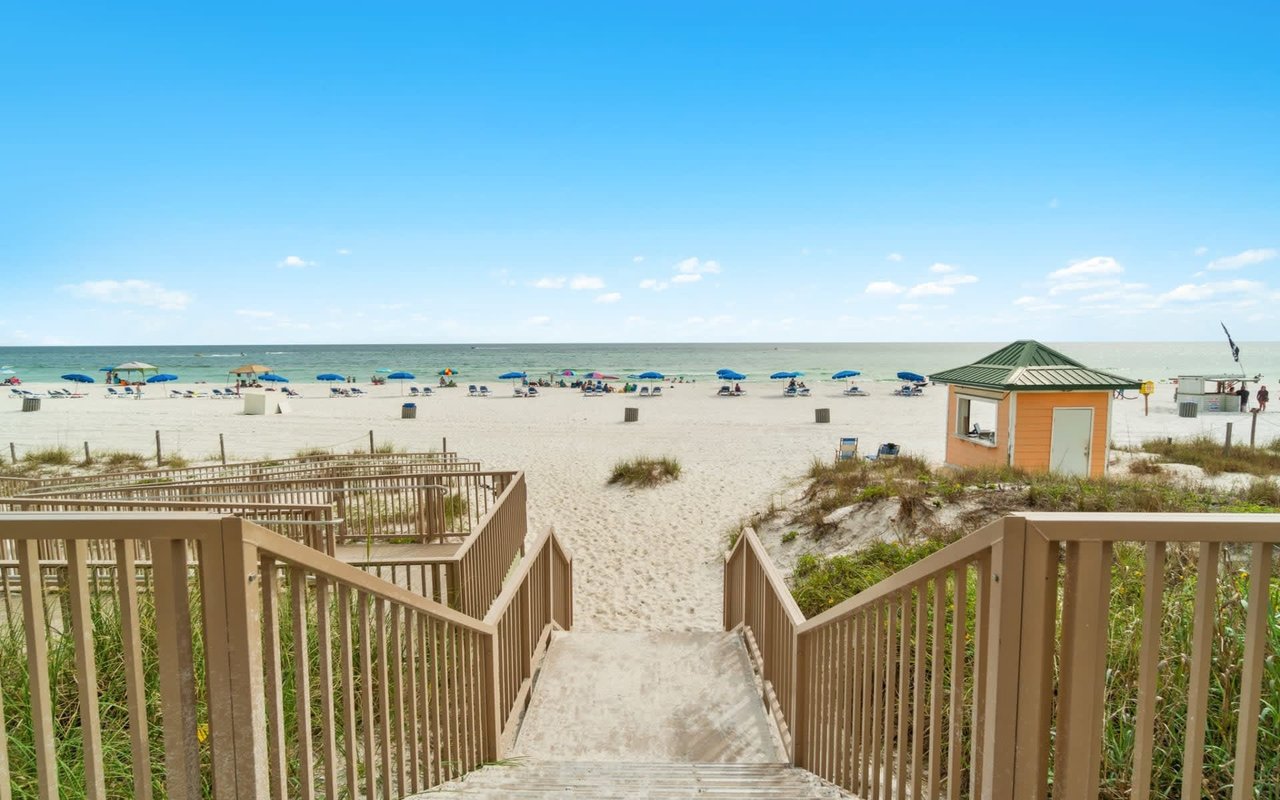 A wooden walkway leads down to a sandy beach with a clear blue ocean.