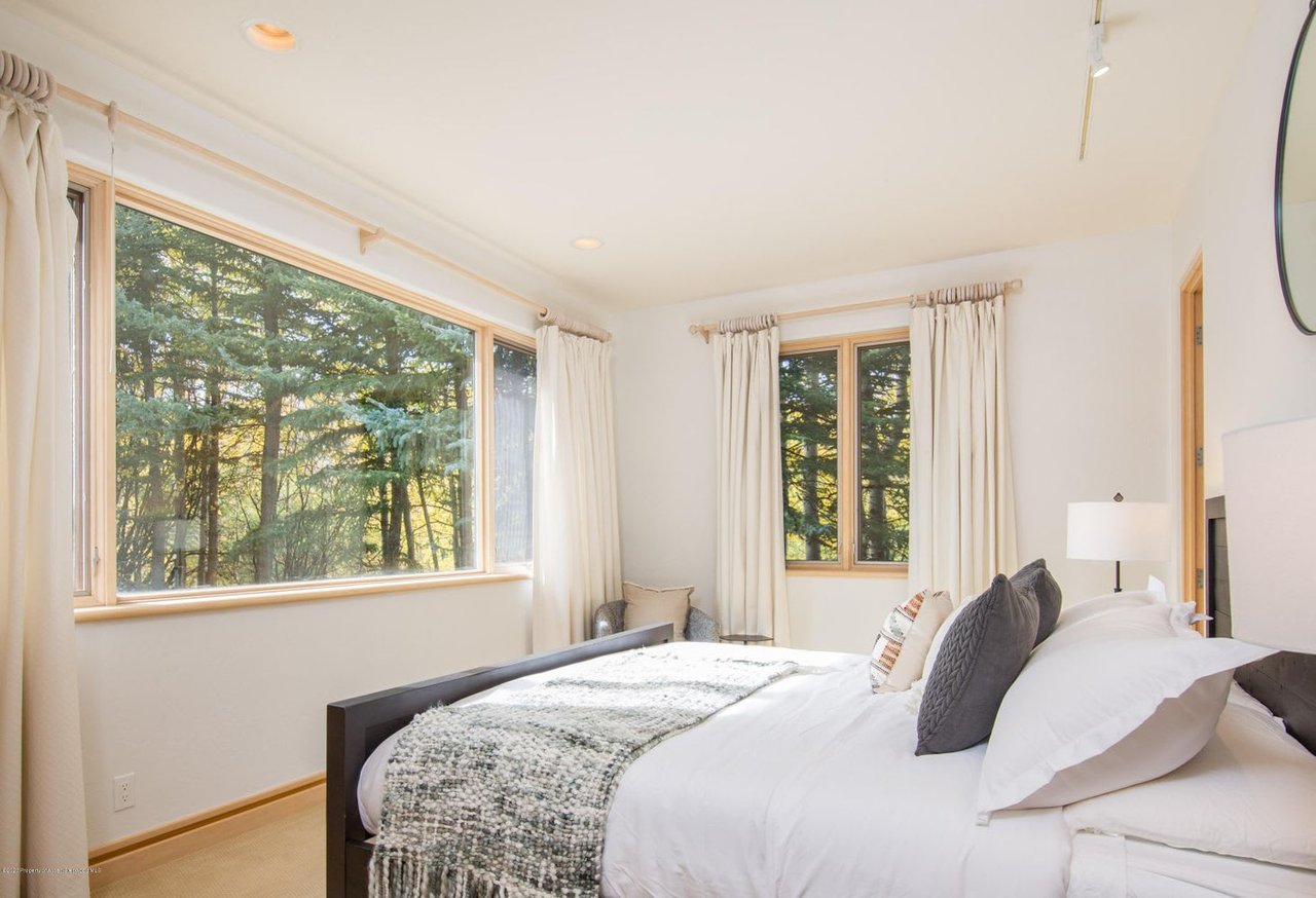 Bedroom with a white bed and large windows overlooking trees.