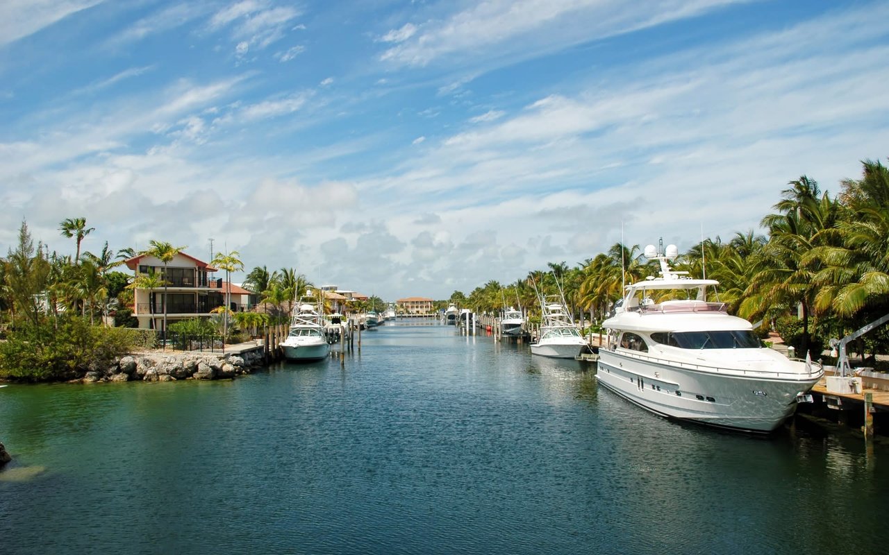 A tranquil canal, possibly in Key Largo, Florida, is lined with luxurious yachts and framed by lush tropical vegetation under a partly cloudy sky.