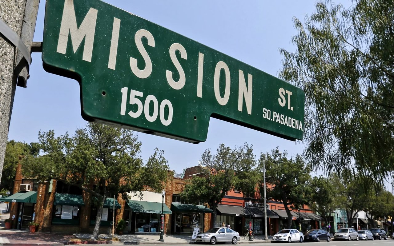 A green street sign hanging from a pole on a city street. The street sign reads as "Mission St." and "So. Pasadena."