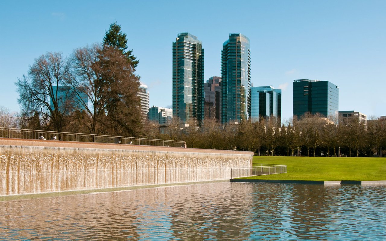 A picturesque view of Bellevue Downtown Park with a large reflecting pool in the foreground, featuring a waterfall spilling over a light-colored wall.