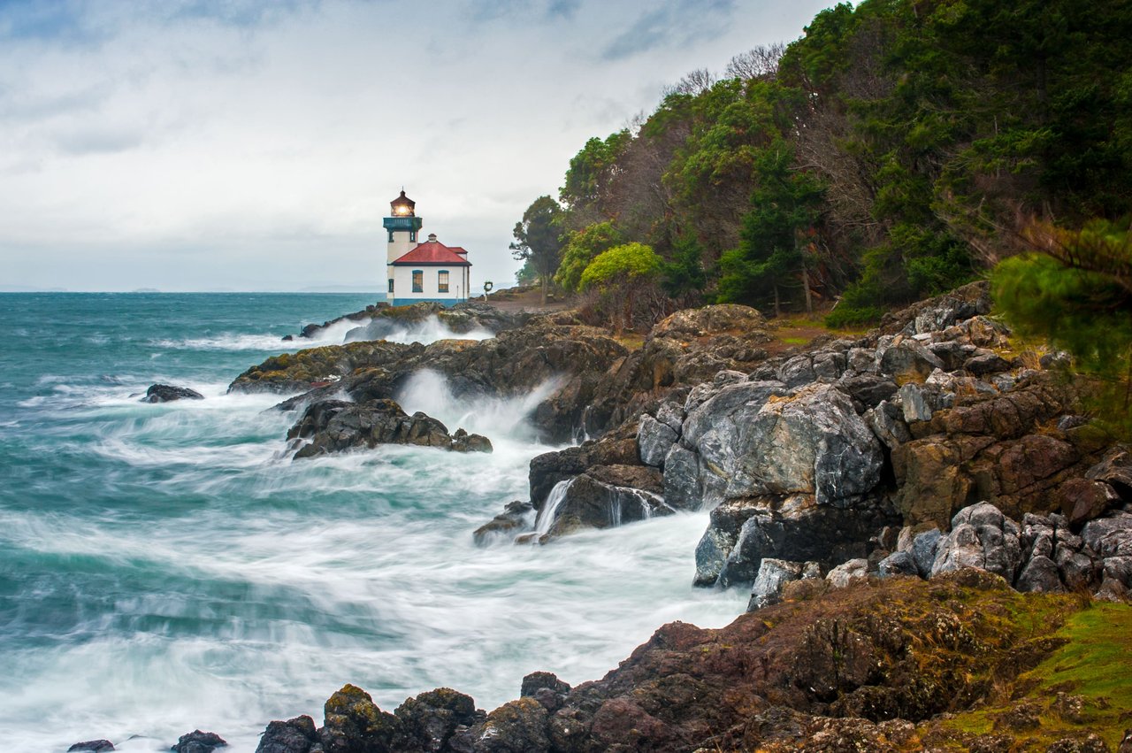 A lighthouse sitting on top of a rocky cliff, with waves crashing against the shore. The lighthouse is white and has a red roof.