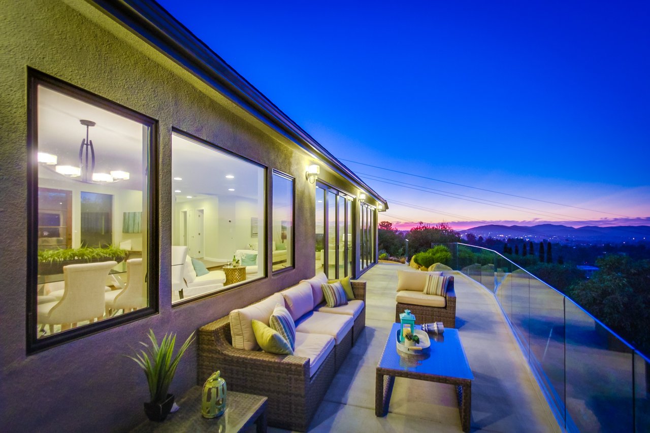 Modern home patio with outdoor seating and glass railing overlooking distant hills at dusk.