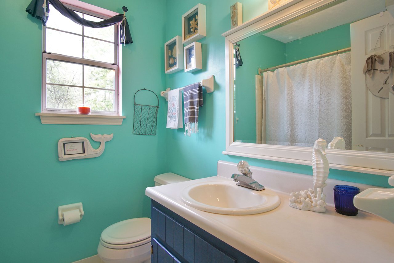 A bathroom with a turquoise color theme. The bathroom has a sink with a mirror, a window, and various bathroom fixtures. The design is bright and cheerful.