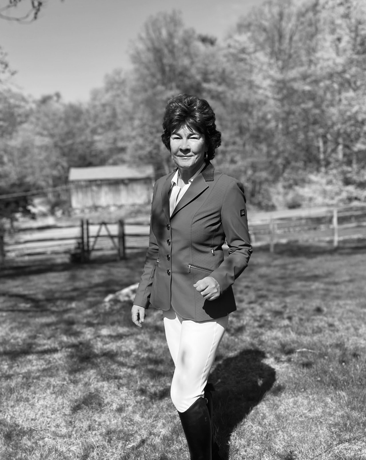 Muffin Dowdle in equestrian attire stands outdoors in a grassy area with trees and a wooden structure in the background, smiling while looking at the camera. The image is in black and white.