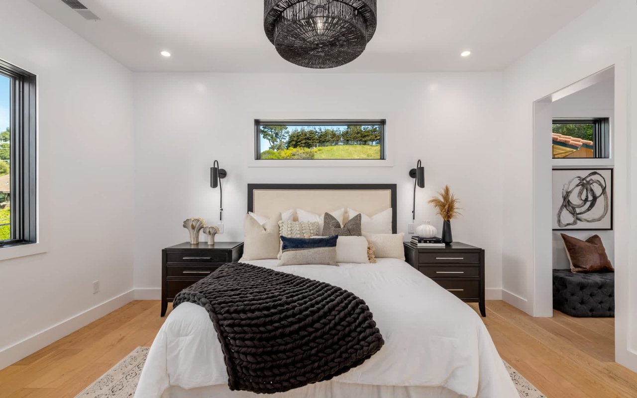 Staged bedroom with a white bed, dark bedside tables, and windows overlooking greenery.