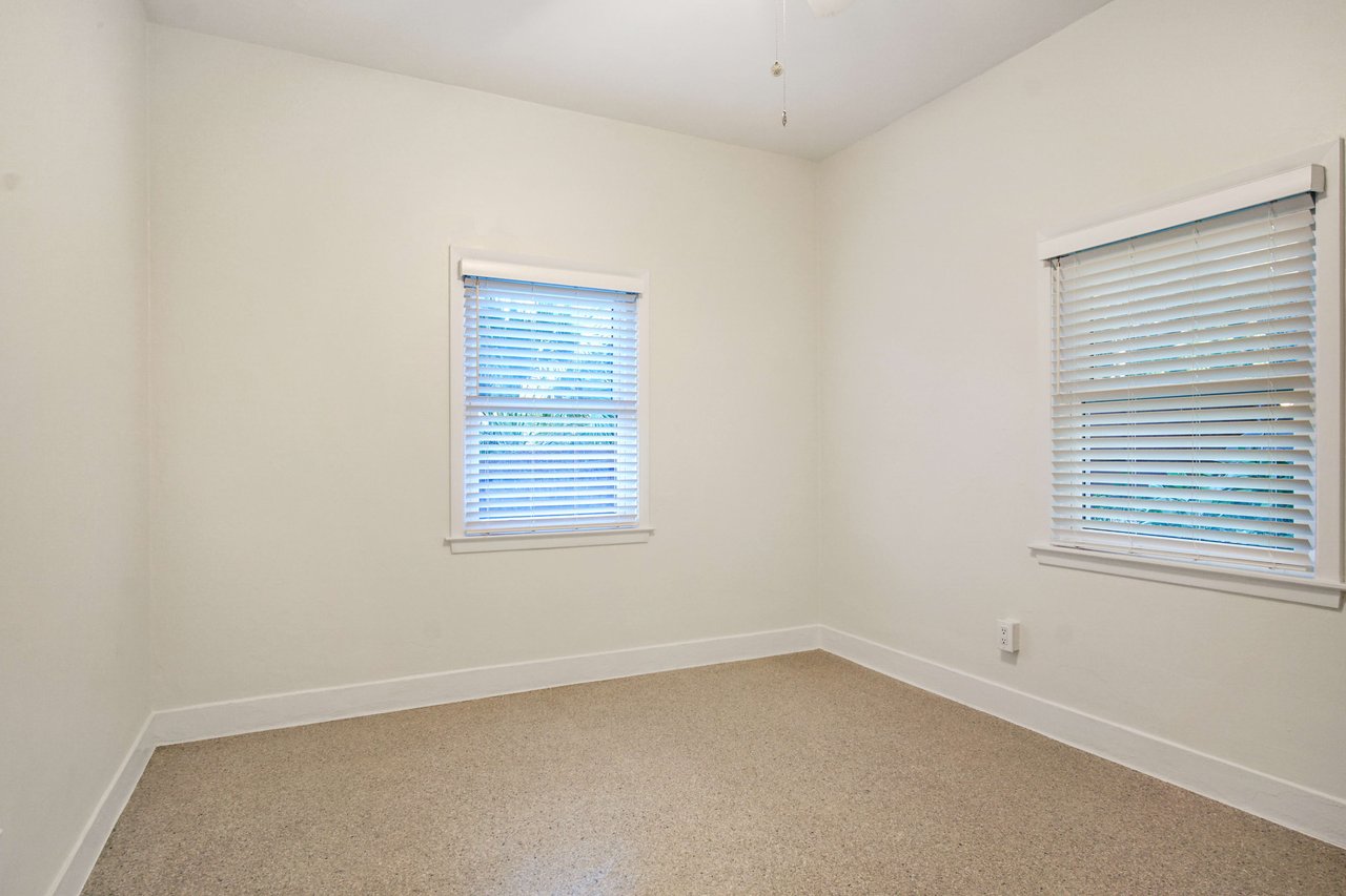 A bedroom with windows on two walls, allowing natural light to fill the space. The room has a simple, clean design.