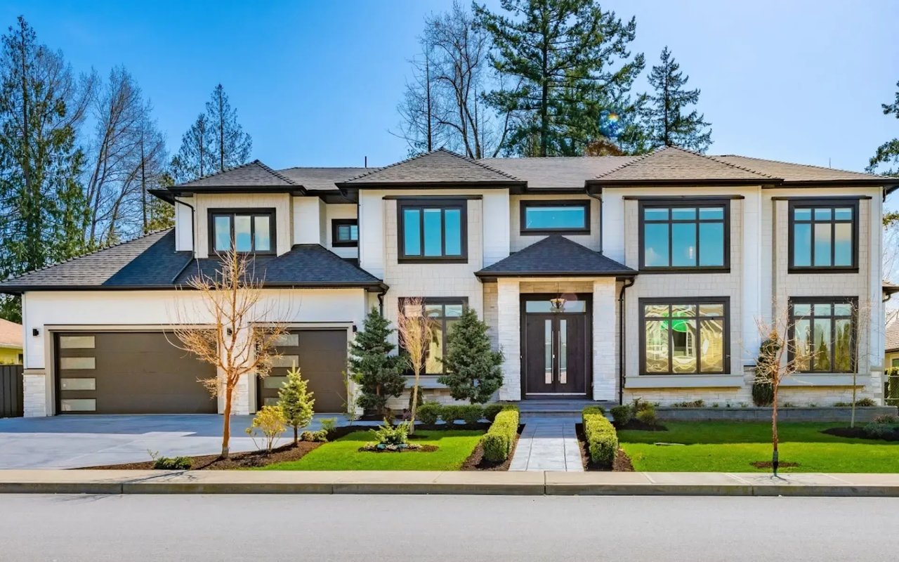 A modern, two-story white house with a brown door and black windows, surrounded by a manicured lawn and tall trees.