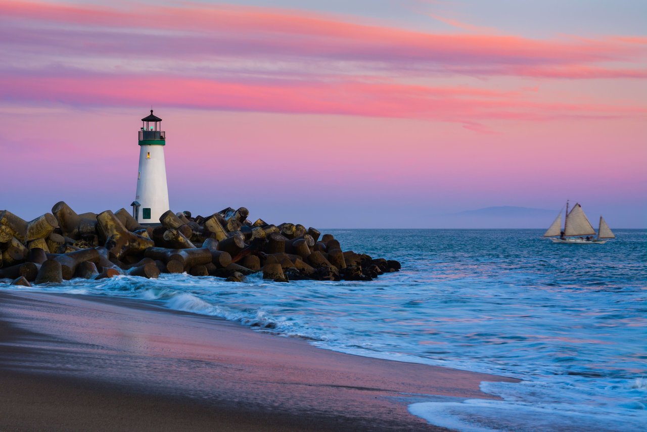 A lighthouse sitting on top of a rocky beach, with a sailboat in the ocean at sunset