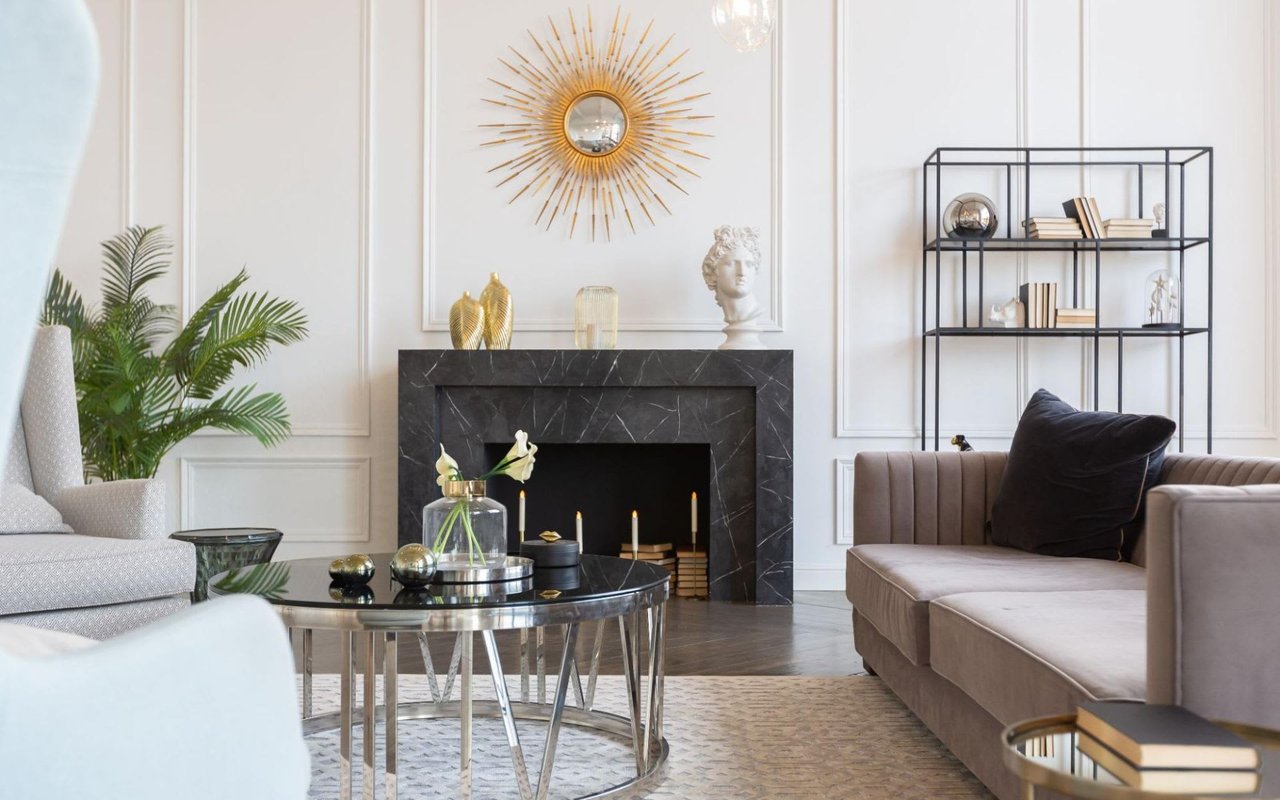 A luxurious living room with a fireplace, a gold sunburst mirror, and a gray sofa, with elegant vases, books, and sculptures.