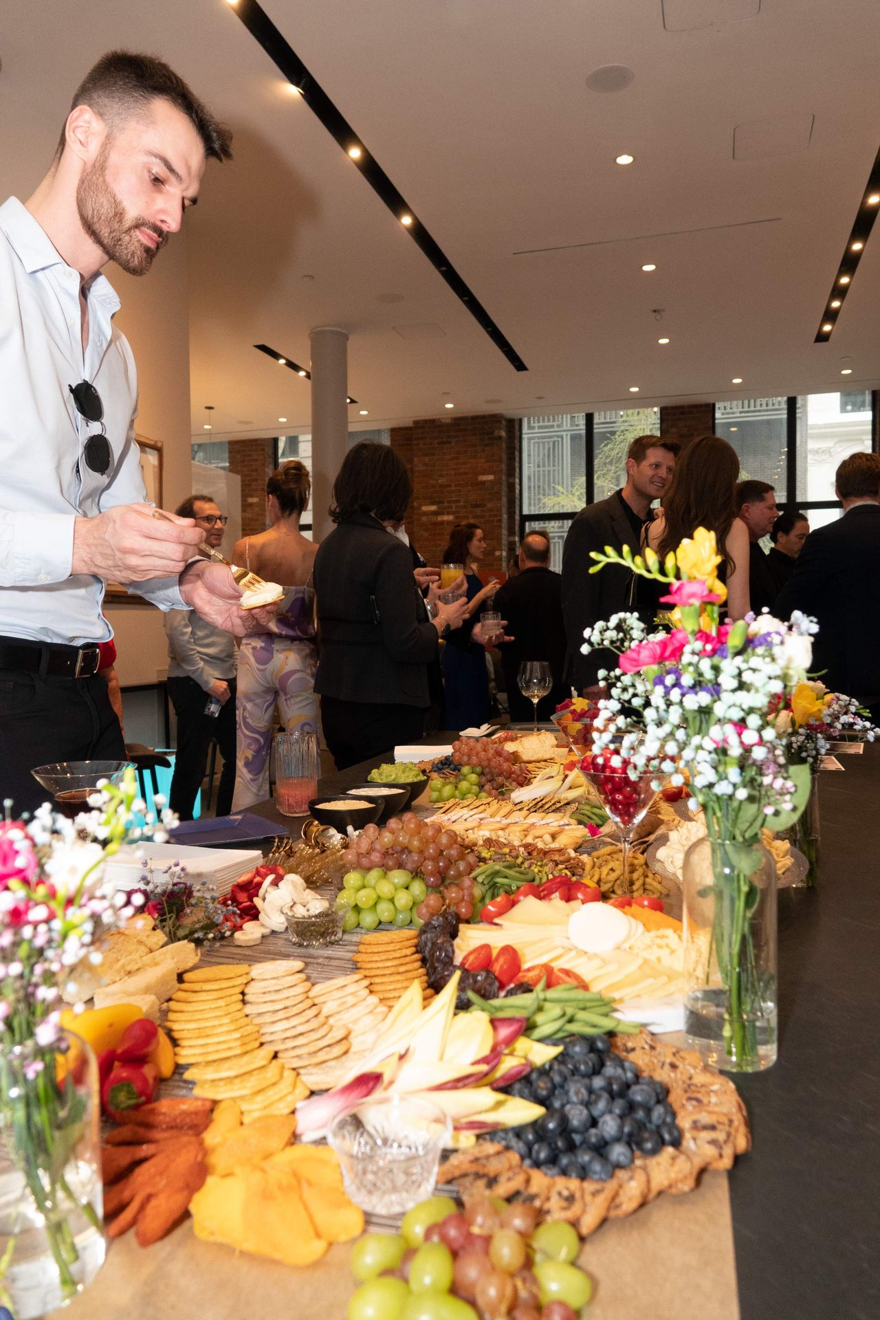 Guests mingle beside a buffet spread with cheeses, crackers, fruit, and flowers.
