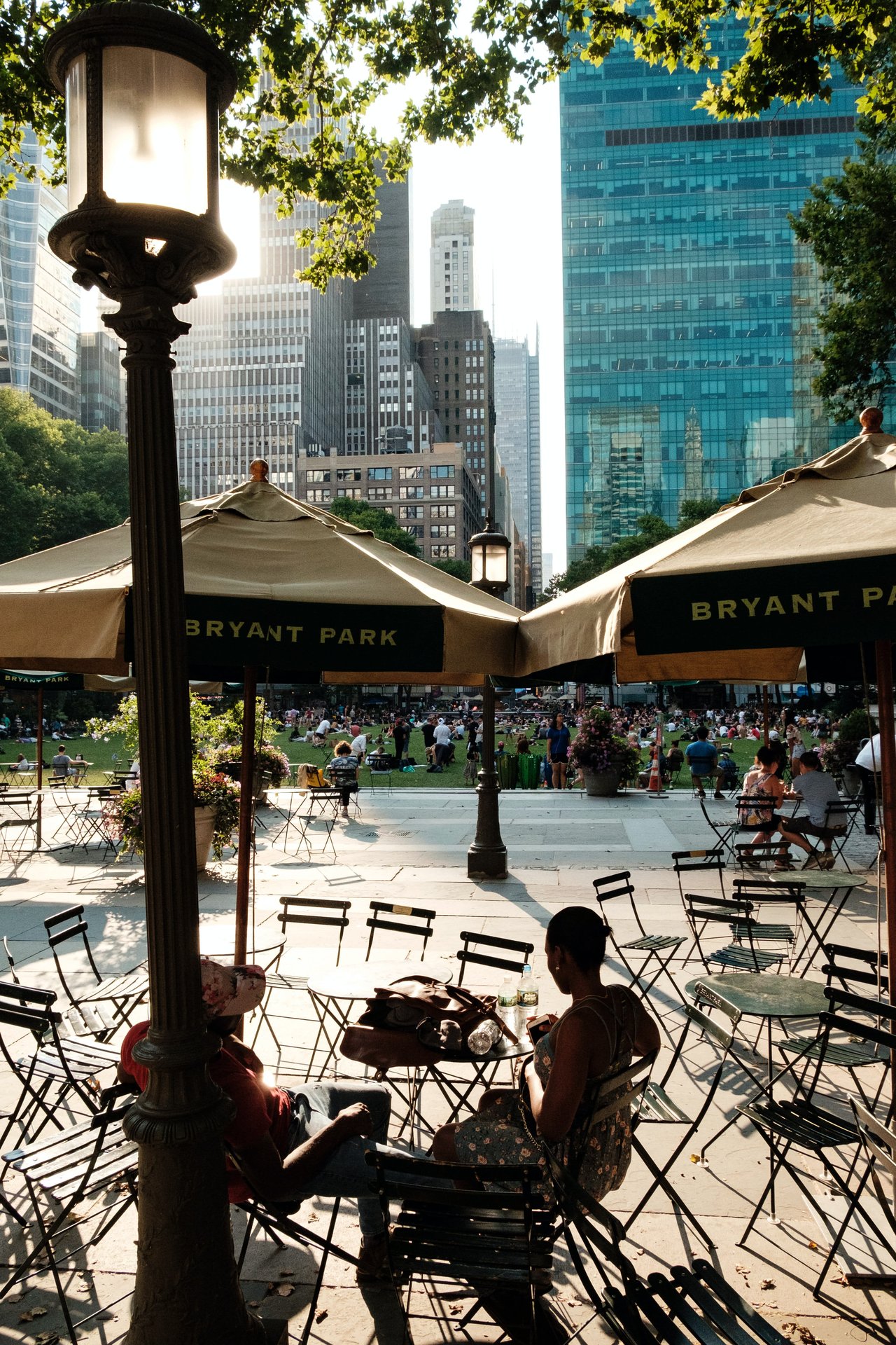 Bryant Park café seating beneath trees, with visitors and Midtown skyscrapers in the background.